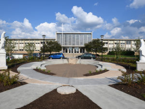 A circular stone and cement plaza in the foreground, surrounded by new landscaping. A statue of a robed woman is on either side of the circle. In the background is the front window-covered facade of a large school in an architecture style common to the 1960s and a large stained glass window above the lobby. Three cars are parked on the road that divides the plaza from the entry to the school. It is a sunny day with blue sky and puffy white and clouds.