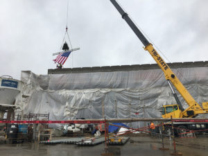 A large yellow crane lifts a ceremonial beam onto the top of Amity Elementary. The beam also holds a small fir tree, and an American flag hangs from the bottom of the beam. "DANGER" ribbons tape off the area in the foreground, and an assortment of construction supplies and equipment can be seen across the area.