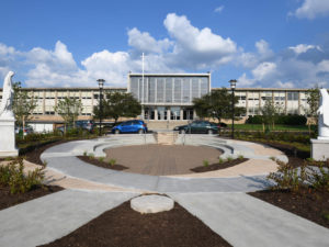 A circular stone and cement plaza in the foreground, surrounded by new landscaping. A statue of a robed woman is on either side of the circle. In the background is the front window-covered facade of a large school in an architecture style common to the 1960s and a large stained glass window above the lobby. Three cars are parked on the road that divides the plaza from the entry to the school. It is a sunny day with blue sky and puffy white and clouds.