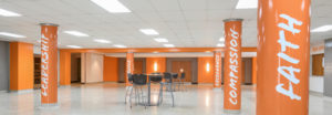 The lobby outside a school auditorium. Five columns are painted bright orange, and have the school values painted on them. From left to right, the columns read, "Leadership," "Service," "Excellence," "Compassion," "Faith." There are two bistro tables in the middle with four black chairs at each of them. The box office is to the far right. The floor is a cool gray, the walls a cheery orange, and a few accent walls are gray or white. Fluorescent lighting is on the ceiling.