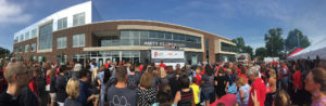A panorama shot of a large crowd gathered in the parking lot of a school on a sunny day with blue sky. The crowd wears a lot of red and black, and faces away from the camera. They are looking at a scaffolding that stands in front of a new elementary school. There are four adults standing at the top of the scaffolding.