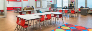 A bright and sunny elementary classroom, ready for the school year. The desks are arranged in clusters. The chairs are a cheerful red, and there are brightly colored bins and books on the shelves along the wall. The door is glass, and an extra bit of glass wall next to the door lets in extra light.