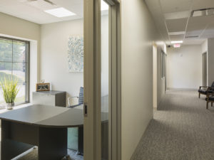 Carpeted hallway of office area. An ajar door allows a view of a well-lit office to the left. Neutral tones, clean lines, and lots of light.